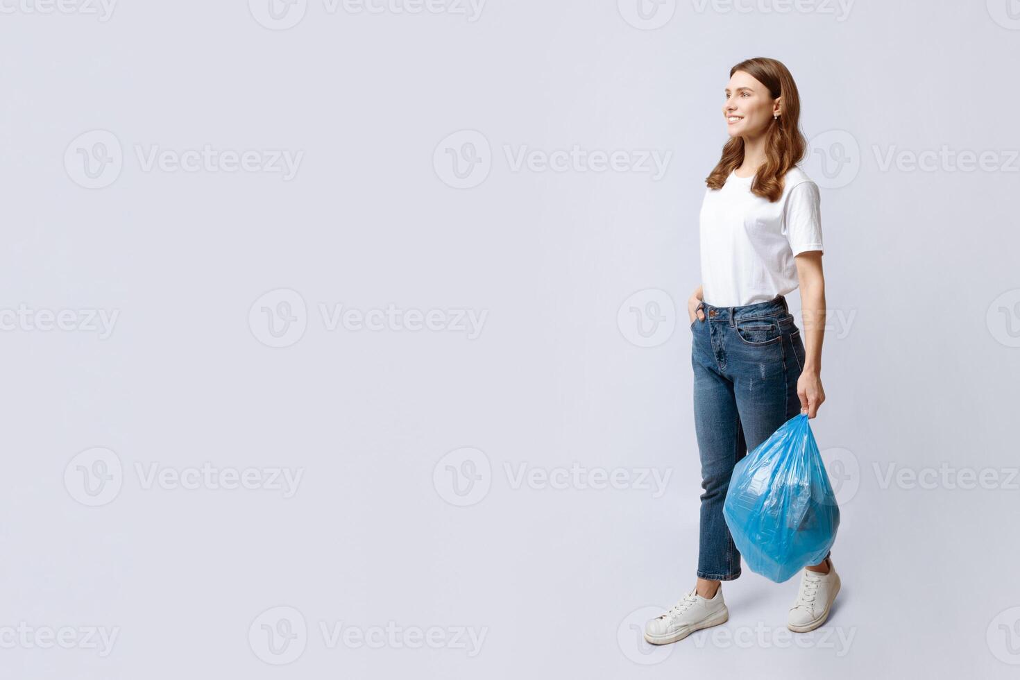 Smiling young housewife walking with plastic bag of household garbage over grey background in studio, positive millennial lady going to throw domestic waste, full-length shot, copy space photo