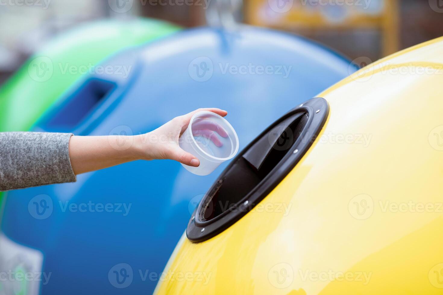 A lady hand places a plastic cup into a recycling bin in a vibrant, eco-friendly setting. The background features colorful bins promoting sustainable practices. photo