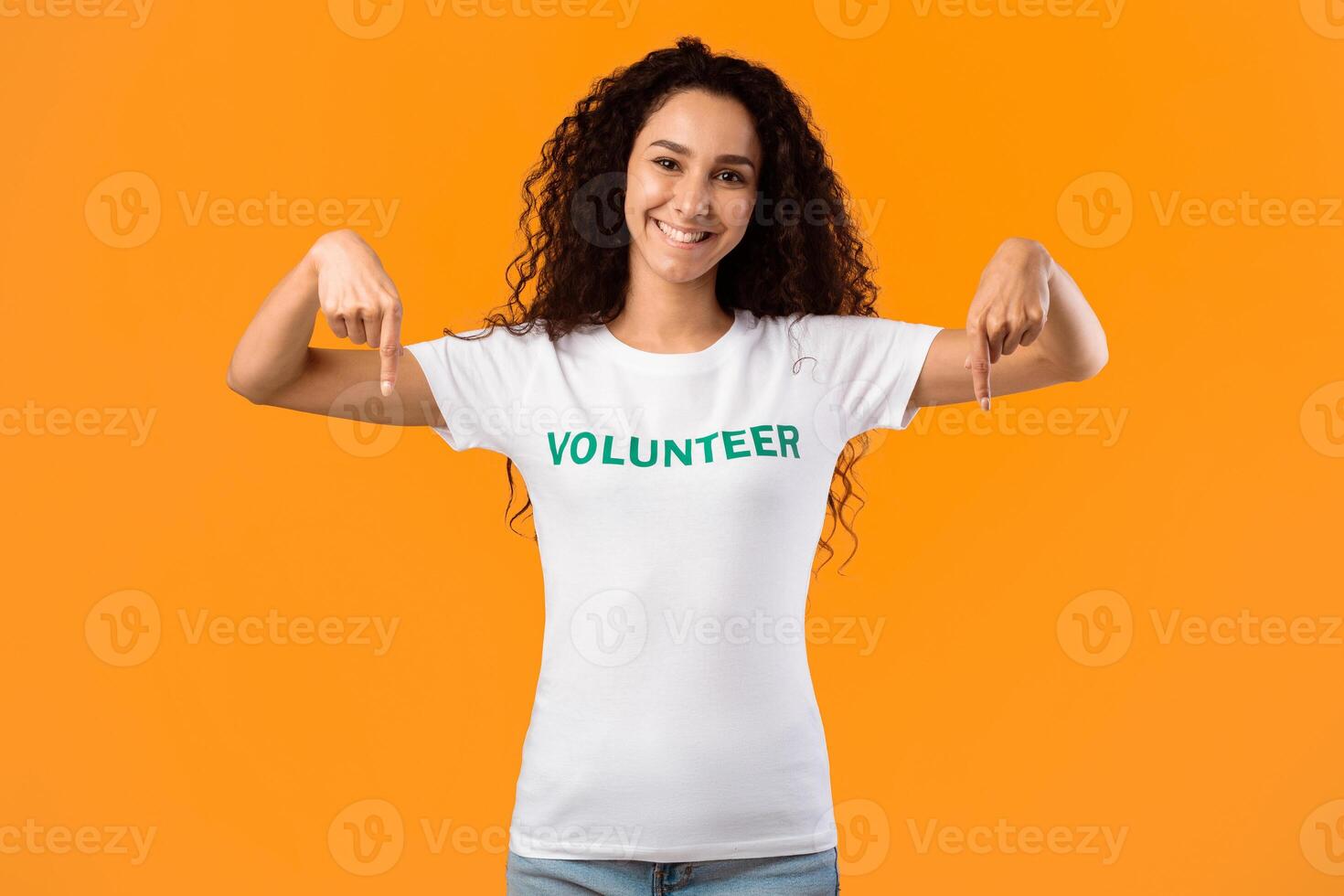 Cheerful Volunteer Lady Pointing Fingers Down Smiling To Camera Posing Standing Over Yellow Studio Background. Activist Advertising Something. Look There, Volunteering Advertisement photo