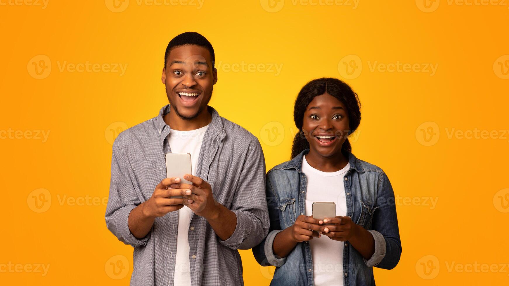 Amazed black man and woman using smartphones over yellow studio background, emotional african american couple playing online games or checking new app, opening mouth in excitement, copy space photo