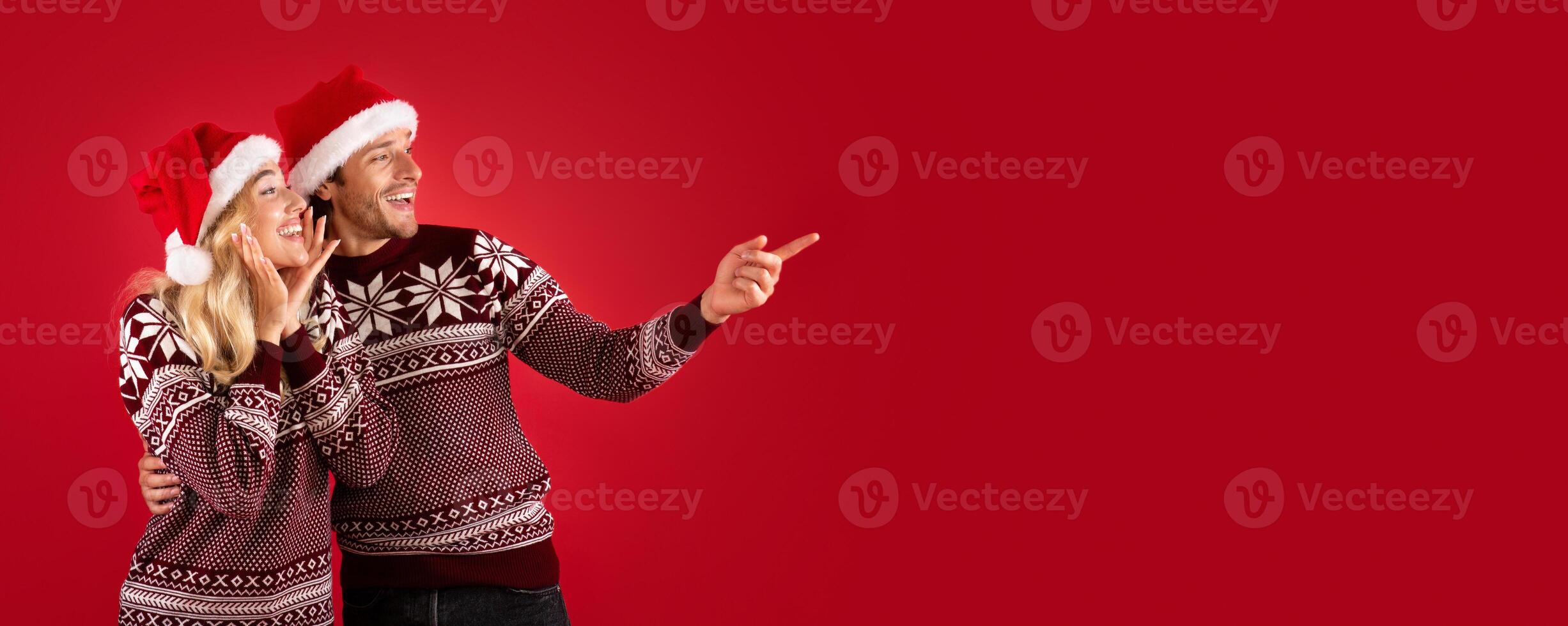 Romantic young couple in Christmas outfits pointing aside at empty space, red background. Panorama photo