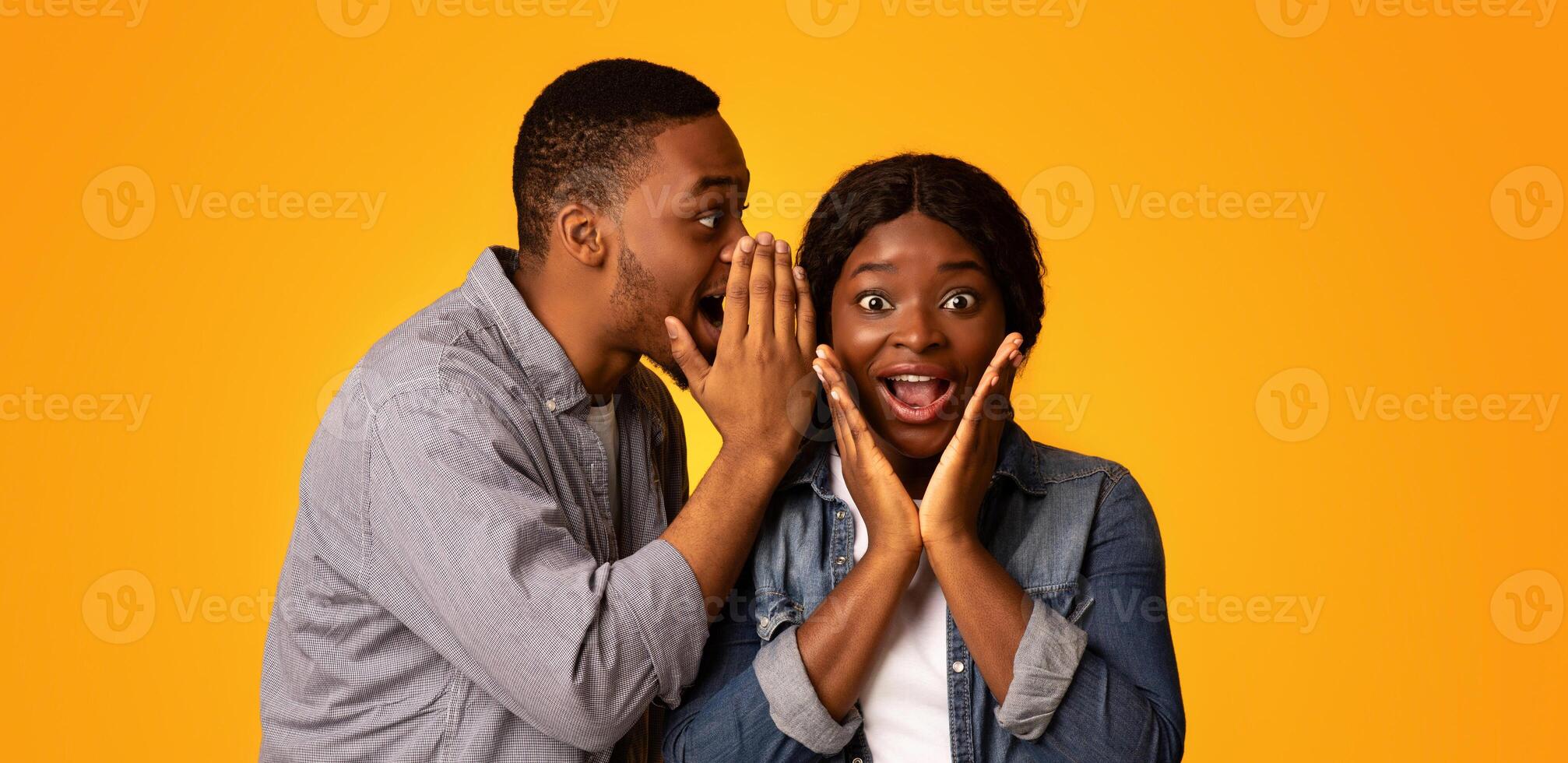 Sharing Secret. Black Guy Gossiping With His Excited Girlfriend, Surprised African American Woman Opening Mouth In Amazement While Standing Together Over Yellow Studio Background, Copy Space photo