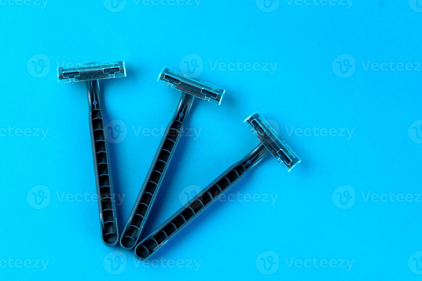 Three disposable razors with black handles and transparent blade covers on a bright blue background. Minimalist shaving tools, hygiene, and personal care concept. photo