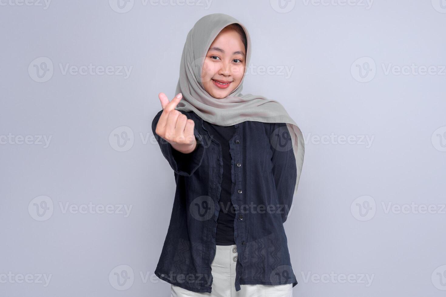 Confident young Indonesian woman in a grey hijab and black blouse smiling while making a Korean finger heart gesture with her hand, standing against a plain studio background. photo