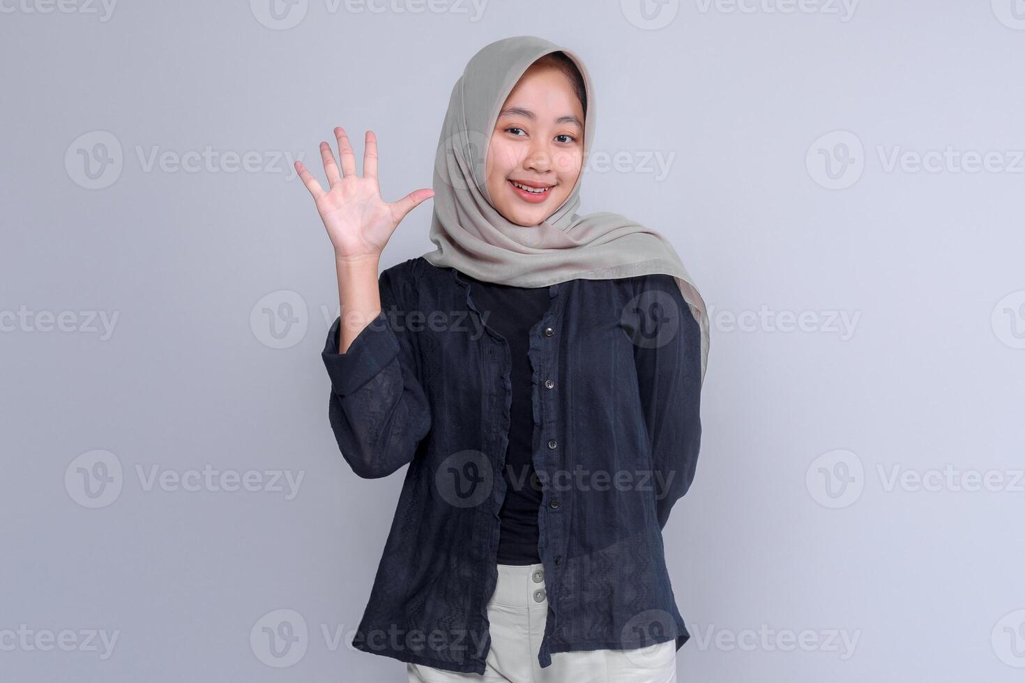 A cheerful young Indonesian woman in a light grey hijab and dark blouse smiles warmly while waving her hand, standing confidently against a plain light grey background. photo