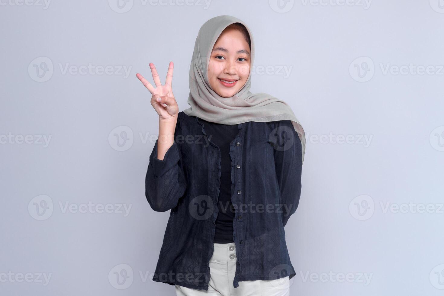 Portrait of a cheerful young Asian woman wearing a light grey hijab and black blouse, smiling confidently while showing three fingers gesture against a plain background. photo