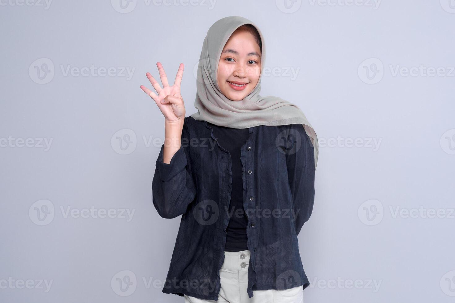 Portrait of a smiling young Indonesian woman in a hijab, showing four fingers with her right hand. She stands confidently against a plain grey background. photo