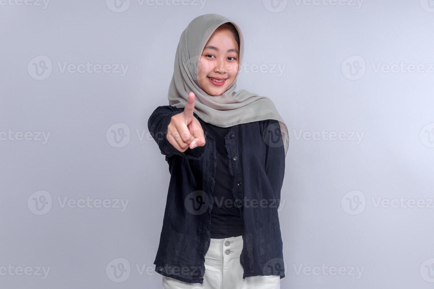 Smiling young Asian woman wearing a hijab, dressed in a black shirt, pointing forward with her index finger. The image conveys confidence, positivity, and an assertive attitude, ideal for branding. photo