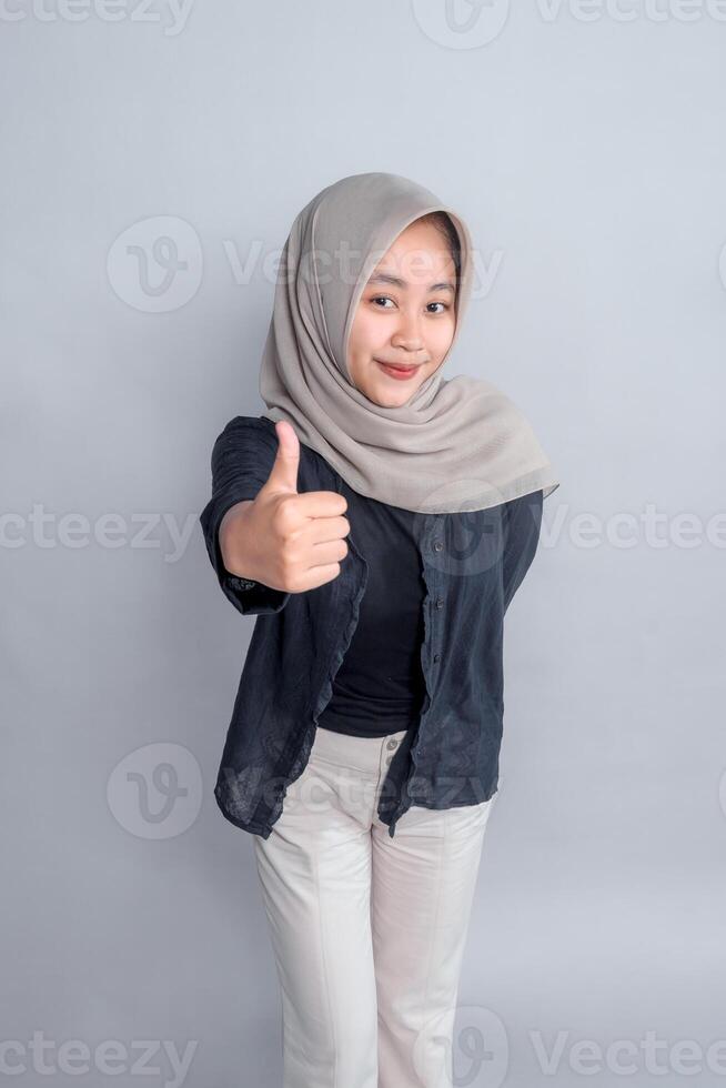Confident young Asian woman wearing a beige hijab and black outfit giving a thumbs-up gesture with a smile, standing against a plain grey background, studio portrait style. photo