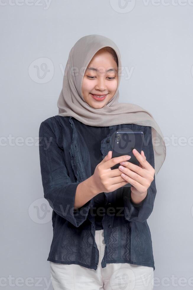 Smiling young Asian woman wearing a beige hijab and dark outfit looking at her smartphone with both hands, standing against grey background, representing digital lifestyle concept. photo