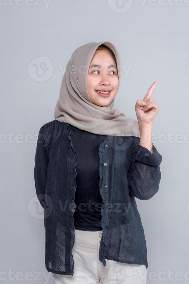 Young Asian woman in beige hijab smiling while pointing up with index finger, wearing dark shirt and cream pants, standing against grey background, isolated. photo