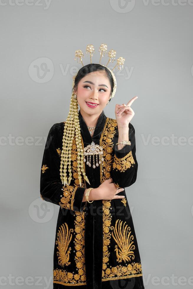 Smiling Asian woman in traditional Javanese dress pointing upward with joyful expression, wearing intricate accessories and elegant kebaya, ideal for cultural and advertising concepts. photo