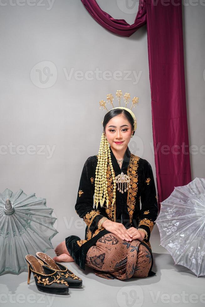 Beautiful Asian woman in traditional Javanese attire smiles calmly while sitting on the floor. Her soft gaze and serene facial expression radiate grace, beauty, and cultural elegance. photo