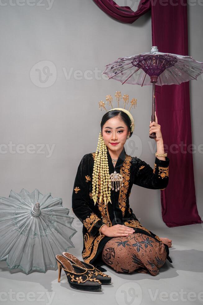 Beautiful Asian woman in traditional Javanese dress sits gracefully, holding a parasol with a gentle smile and confident expression, ideal for cultural and commercial visuals. photo