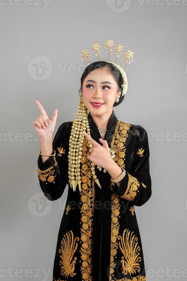 Beautiful young Asian woman wearing traditional Javanese kebaya and intricate golden accessories, smiling confidently while pointing upward with expressive, engaging facial expression. photo