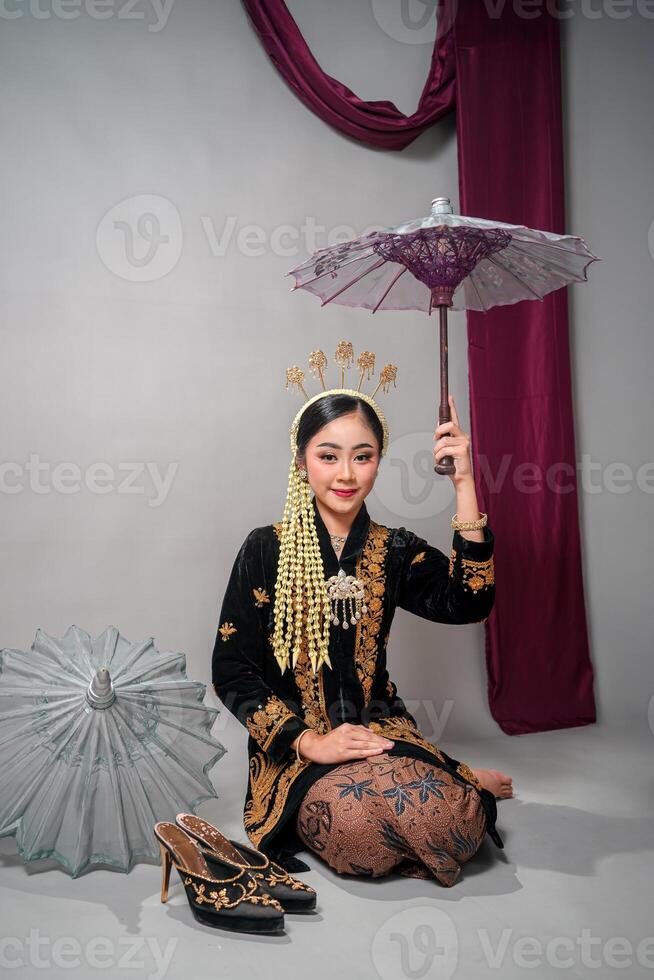 Asian woman in traditional Javanese costume smiles softly while holding a decorative umbrella. Her gentle expression highlights elegance, cultural pride, and classic feminine charm. photo