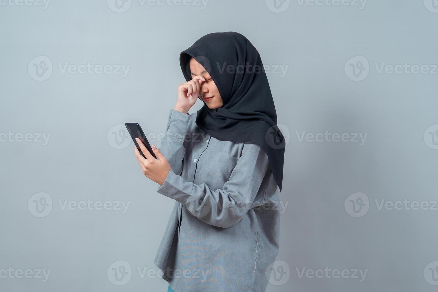 Portrait of a young Muslim woman in a hijab rubbing her tired eyes while holding a smartphone, expressing digital eye strain or screen fatigue in a modern lifestyle context. photo