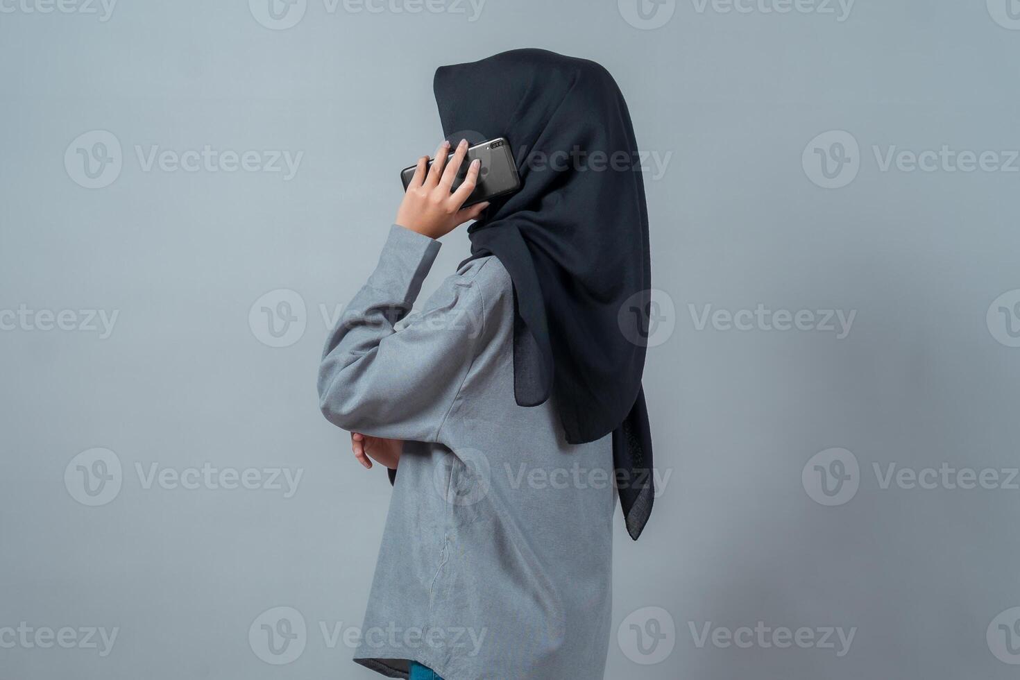 Side view of a young Asian Muslim woman wearing a black hijab and grey shirt, standing against a plain background while making a phone call using a smartphone, appearing calm and focused. photo