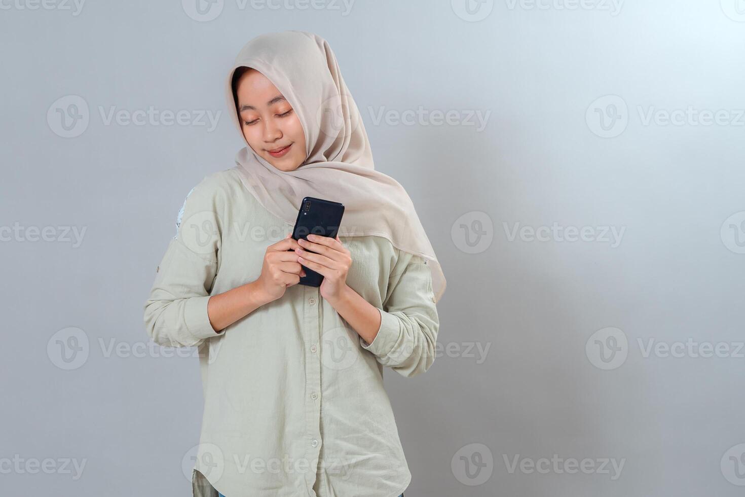 Young Asian Muslim woman wearing hijab holding smartphone with both hands, smiling gently with eyes closed, expressing happiness, affection, or sweet emotional reaction indoors. photo