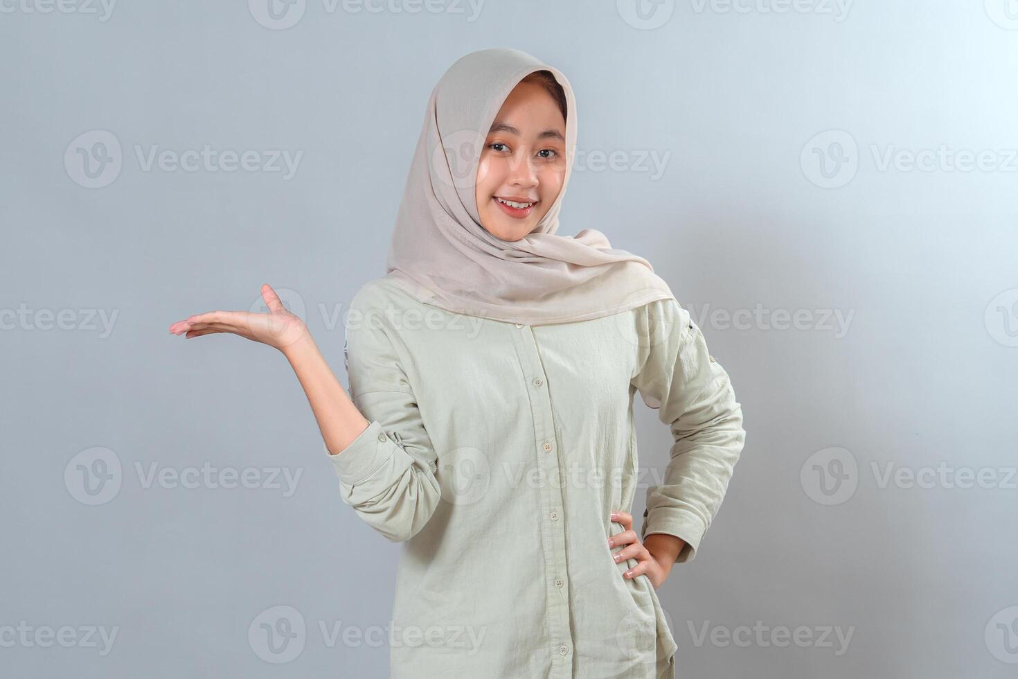 Portrait of a young Muslim woman in hijab smiling warmly while raising one hand in a presenting gesture. Her expression is friendly, confident, and inviting, perfect for advertisement use. photo