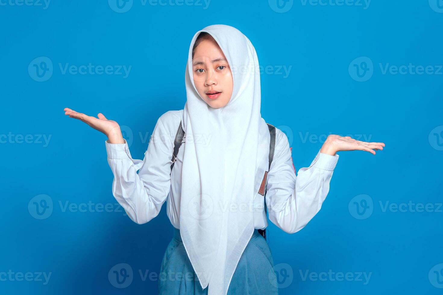 Confused Asian Muslim teenage girl in school uniform with backpack, shrugging with uncertain expression, isolated on blue background, back to school concept. photo