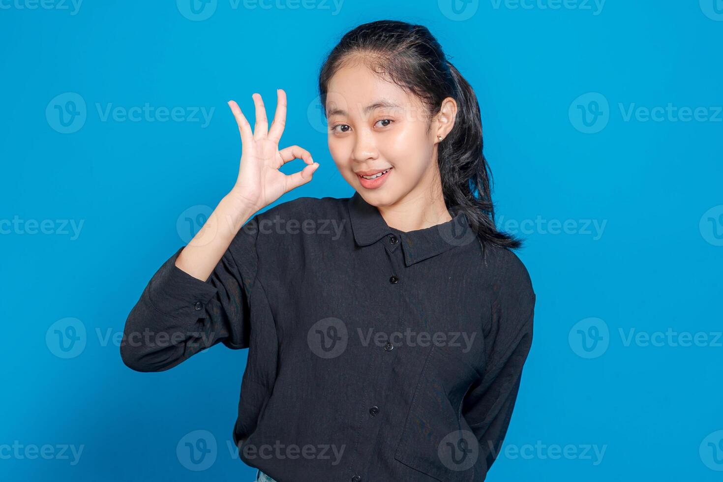 Smiling young Asian woman making an OK hand gesture while looking at the camera. Her friendly and confident facial expression conveys positivity, approval, and satisfaction. photo