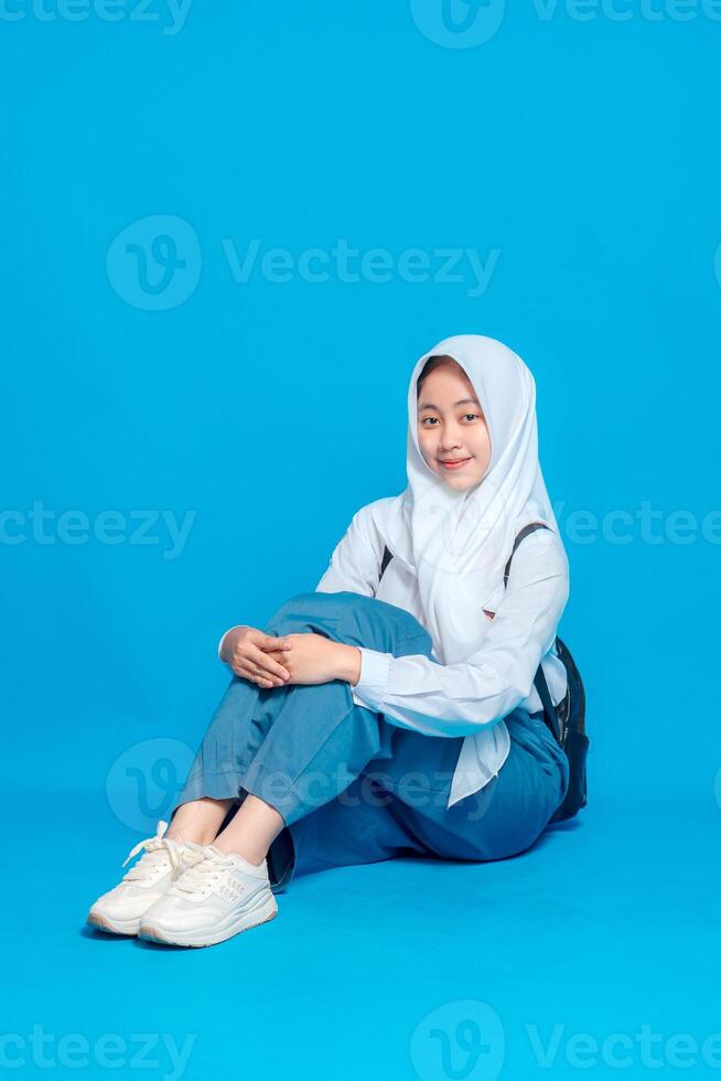Asian Muslim teenage girl student sitting on floor with cheerful smile and relaxed pose, wearing hijab and school uniform, ready to learn, education and back to school concept. photo