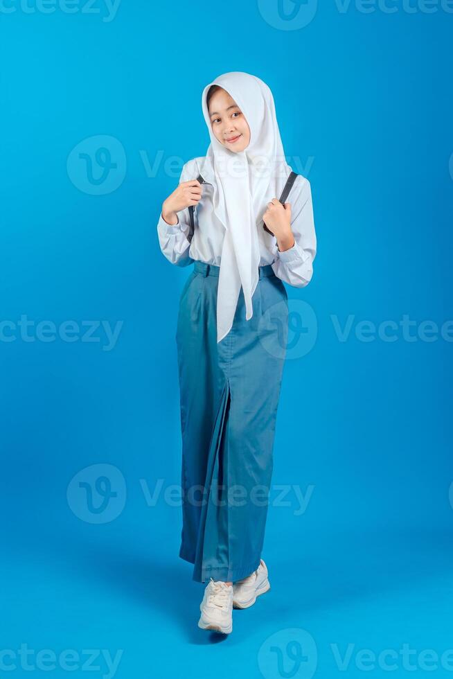 Asian Muslim teenage girl student smiling sweetly with positive expression, wearing hijab and school uniform, carrying backpack, standing confidently, back to school education concept. photo