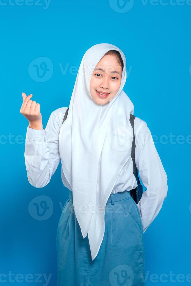 Happy Muslim schoolgirl wearing a white hijab and uniform, making a mini heart hand gesture while smiling sweetly. She looks cheerful and confident on a blue background. photo