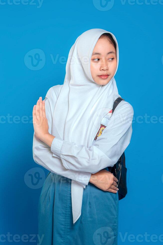 Confident Muslim schoolgirl in uniform and hijab showing stop hand gesture with calm, assertive facial expression. Back to school concept on isolated blue background. photo