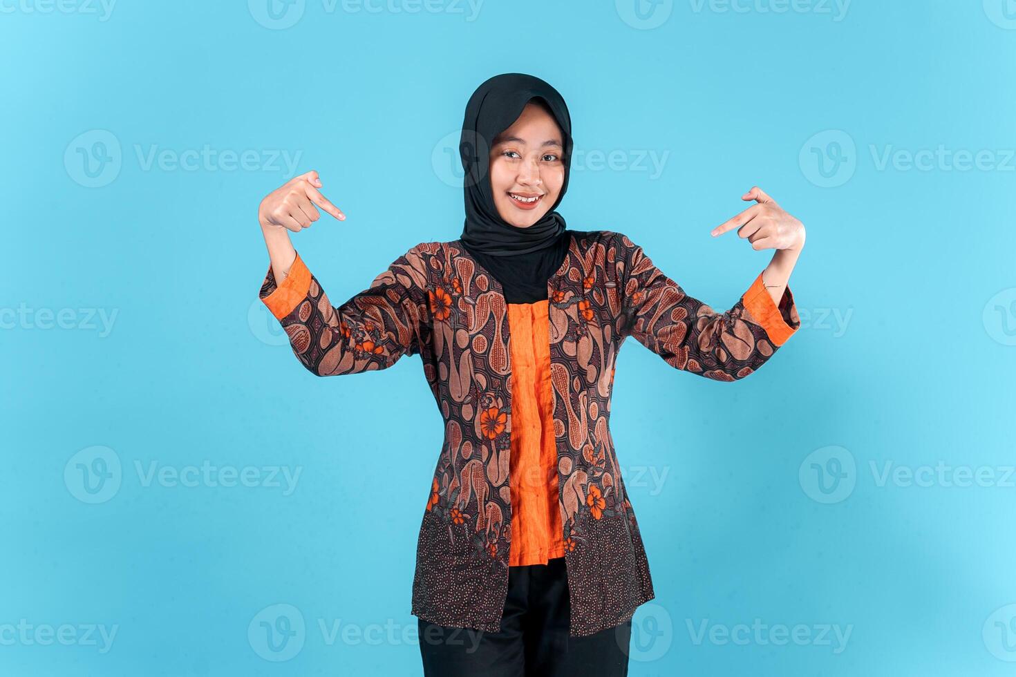 A cheerful young Asian woman in a hijab and batik outfit smiles while confidently pointing both index fingers downward, showcasing a playful and friendly facial expression in studio. photo