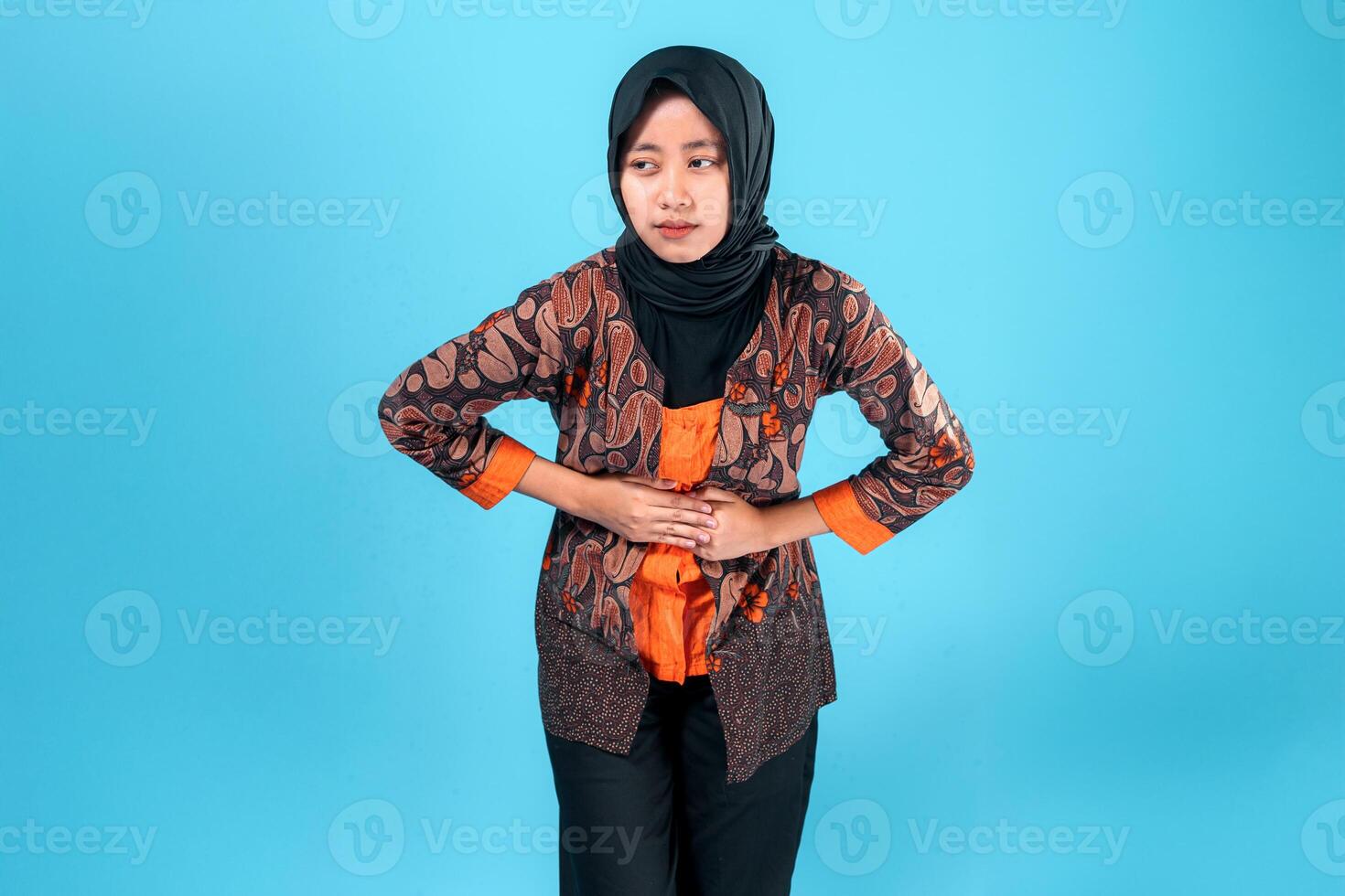 A young Asian woman in a batik outfit and hijab appears to be in discomfort or pain, holding her stomach with both hands while showing a concerned facial expression. photo
