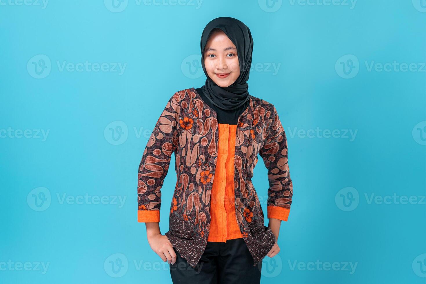 Smiling young Asian woman in traditional batik outfit and hijab posing confidently against blue background, showing friendly and welcoming expression. photo