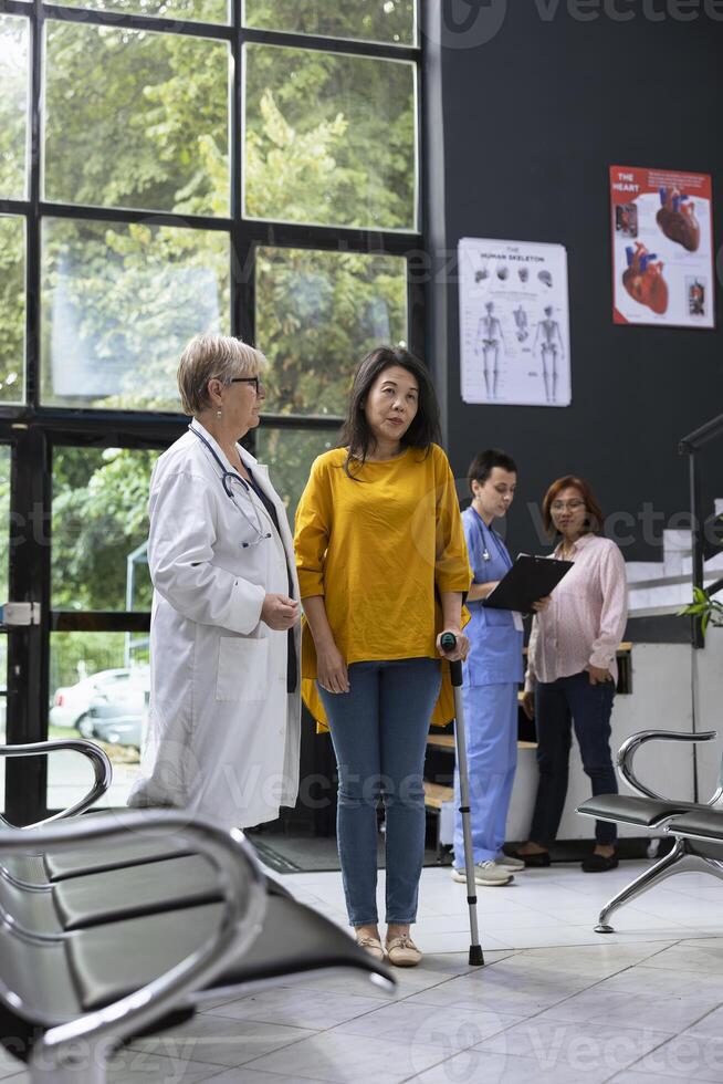 Staff assists asian woman with locomotor problems walk on crutches through a waiting area lobby, highlighting rehabilitation, support and inclusive healthcare services. Healing process. photo