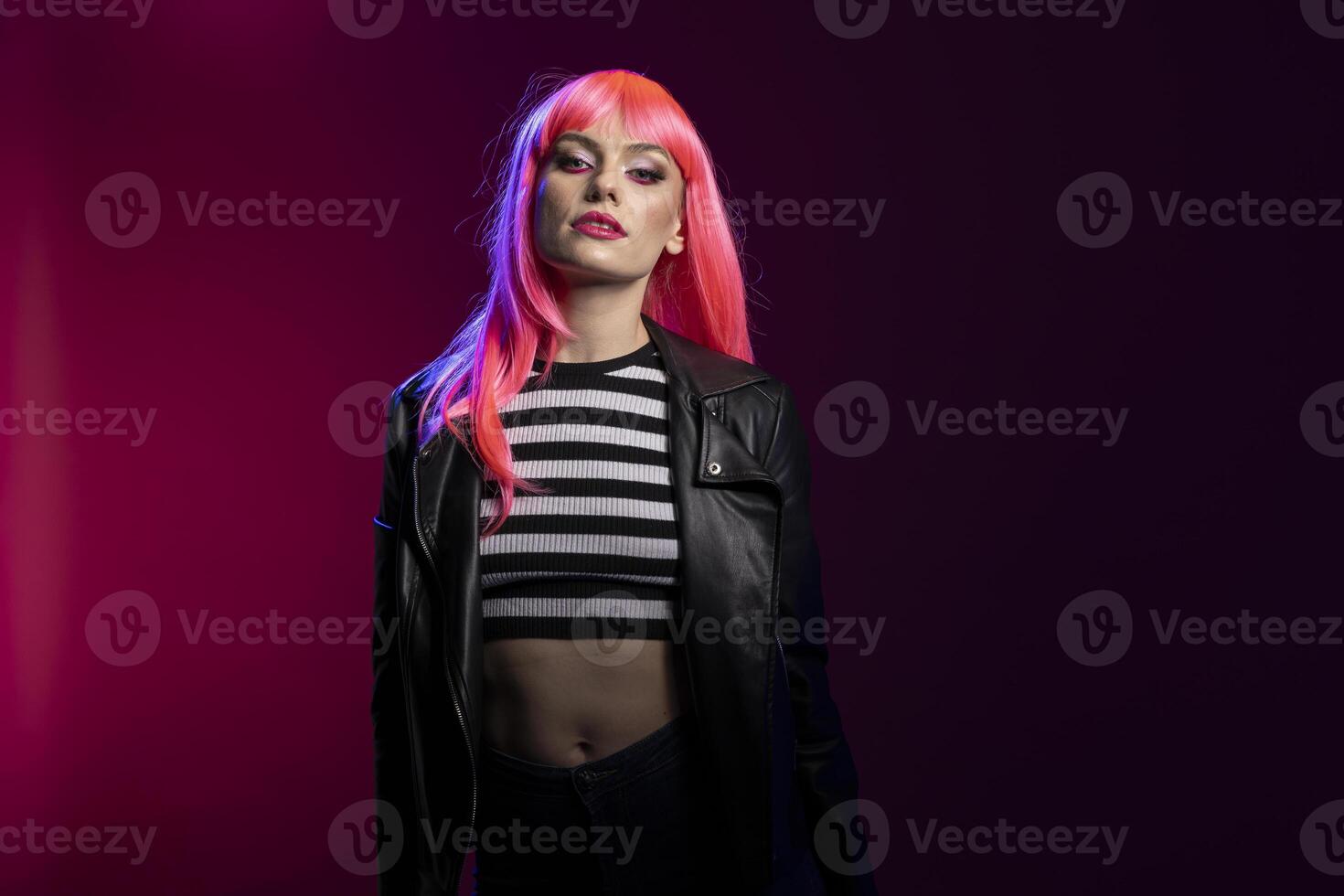 Confident female model throws bold attitude with intense eye contact and edgy styling. Pretty woman with pink hair and striped top pop posing under creative blue and magenta studio lights. photo