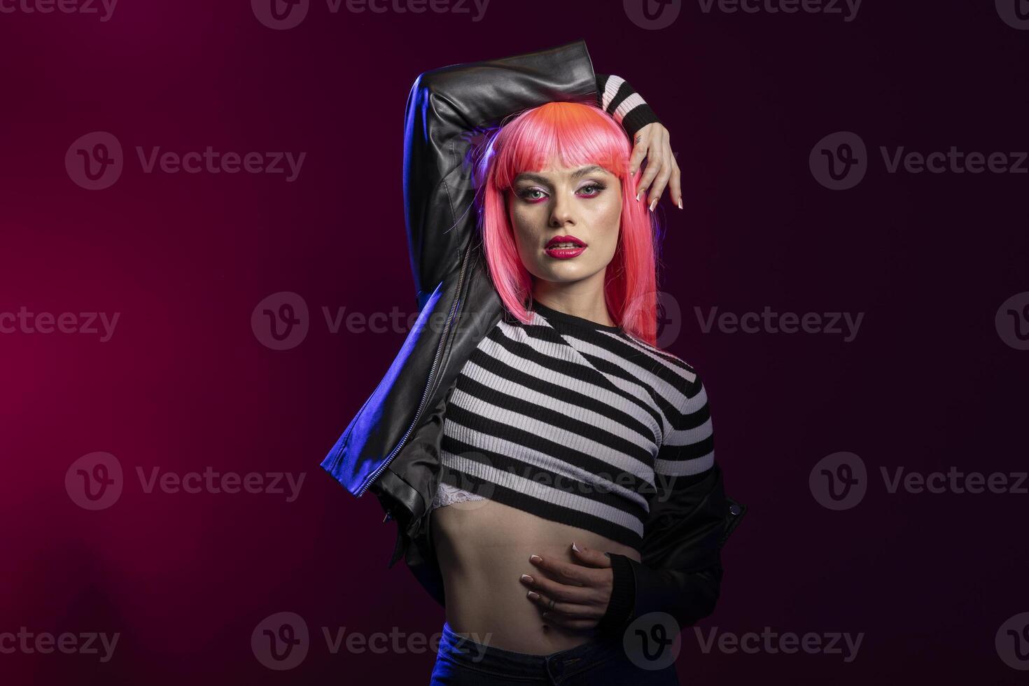 Strong elegant woman projects attitude in studio using bold expression and confident posture. Fierce female model with pink hair, wearing black leather jacket poses against isolated background. photo