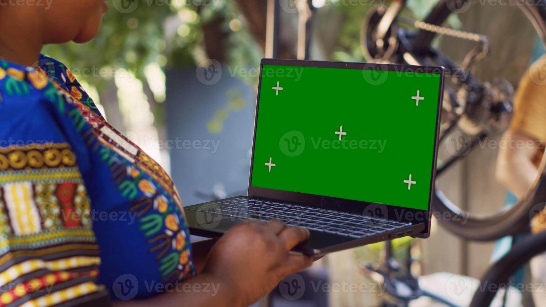Detailed view of african american lady grasps minicomputer with greenscreen display as man in background removes broken wheel. Black woman holds laptop with blank mockup screen for bike maintenance. photo