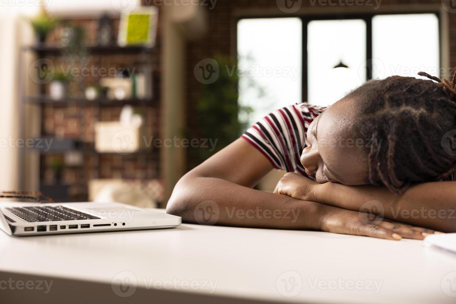 African american female freelancer naps on desk beside laptop in home office, physically drained from working under tight deadline. Woman entrepreneur peacefully sleeping after completing tasks. photo