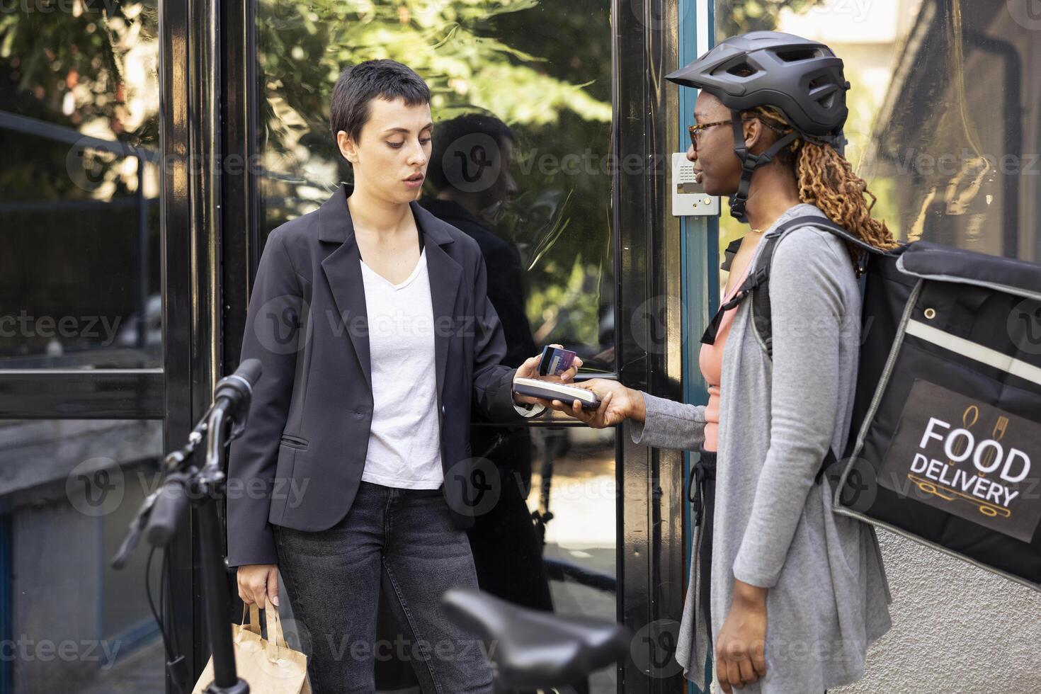 Black female worker accepts electronic payment from customer with a card on pos terminal. Courier ensuring transaction finalizes the purchase of fast food order from restaurant delivery. photo