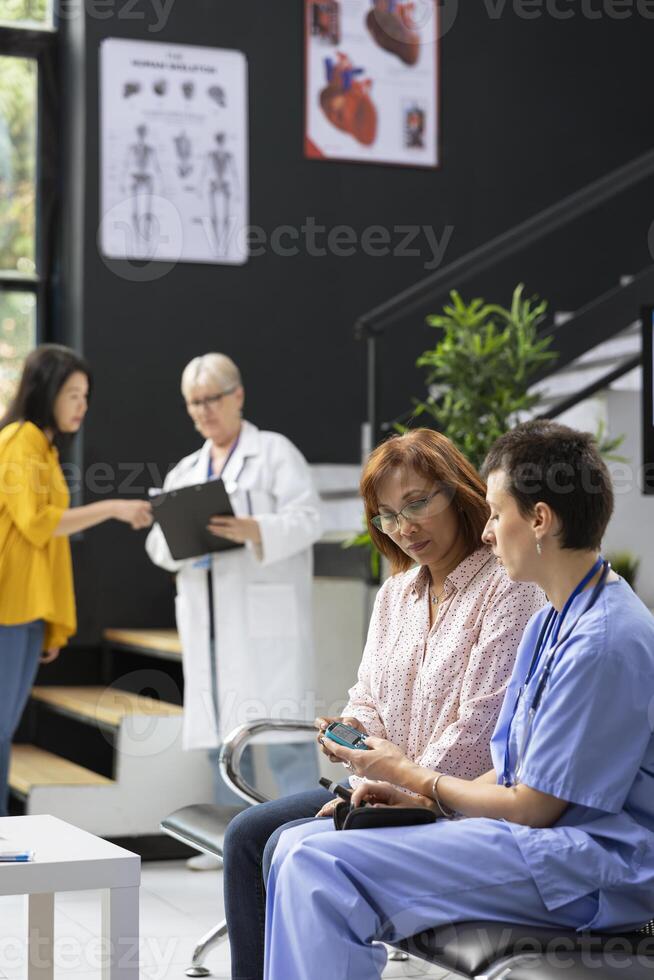 Nurse assisting asian patient in measuring her sugar levels with glucometer for a routine checkup in waiting room. Specialist explains diabetes reading interpretation for glucose levels. photo