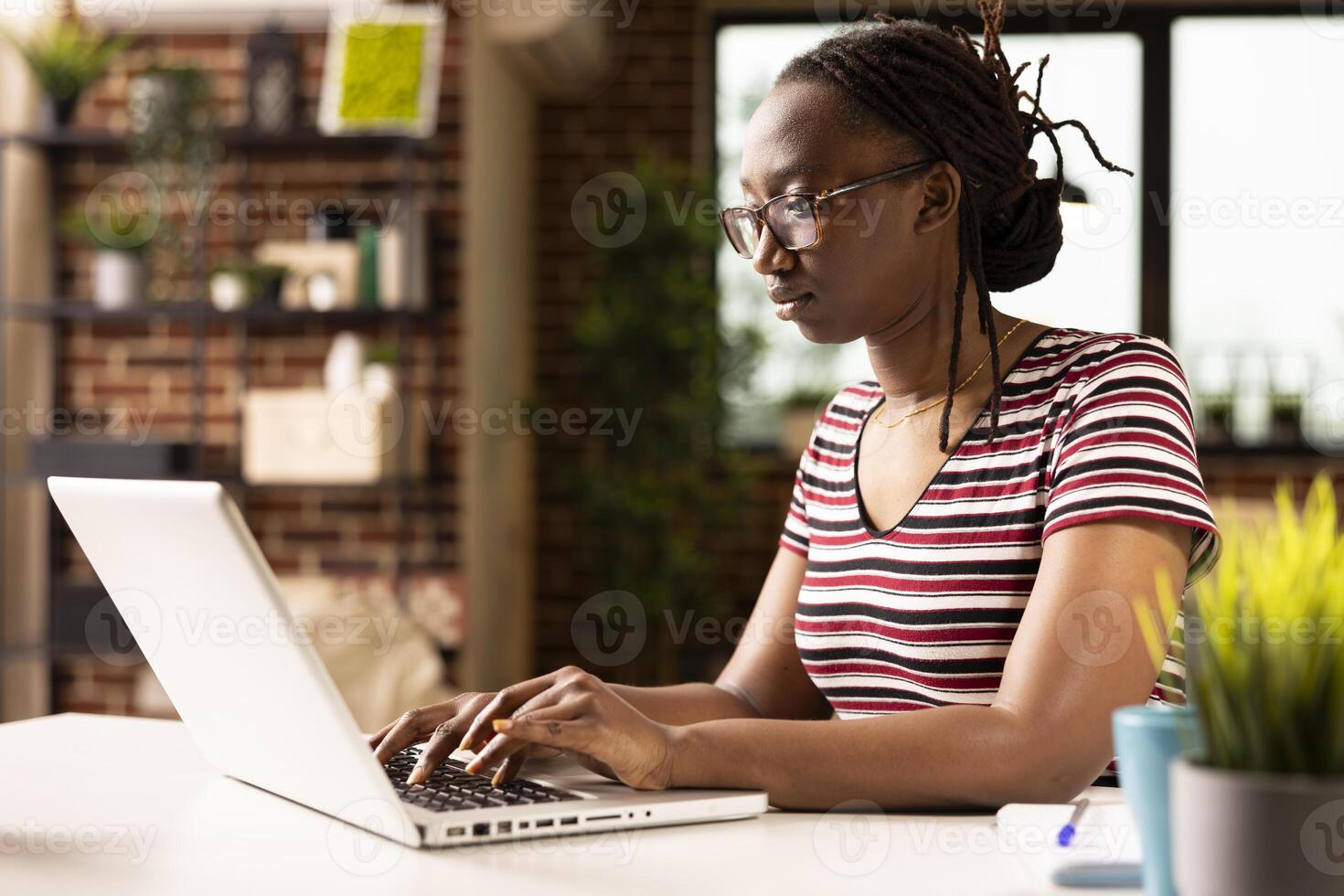 Self employed black woman working on laptop, updating her personal business platform in home office. Focused female manager reviewing digital financial files and managing project tasks. photo