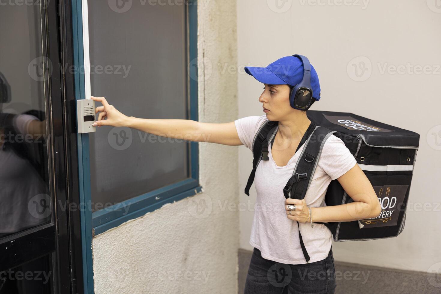 Delivery employee with thermal backpack arrives at the door ringing the bell, approaches a doorway to deliver a takeout meal order. Woman takeaway service courier using the intercom. photo