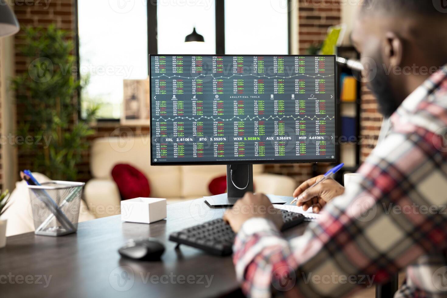 Male entrepreneur writes notes, tracking stock market trends and planning startup investments. Self employed man at home office, analyzing financial data on computer and jotting down ideas on notepad. photo