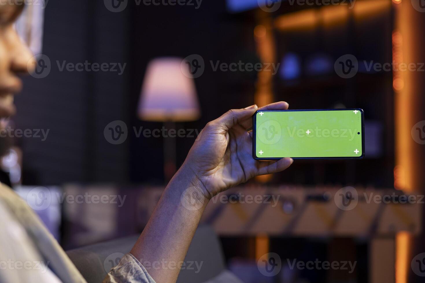 African american male freelancer holding phone with mockup copy space screen, relaxing on sofa in a space with neon lights and smart tv setup. Young person feels casual at his home. photo