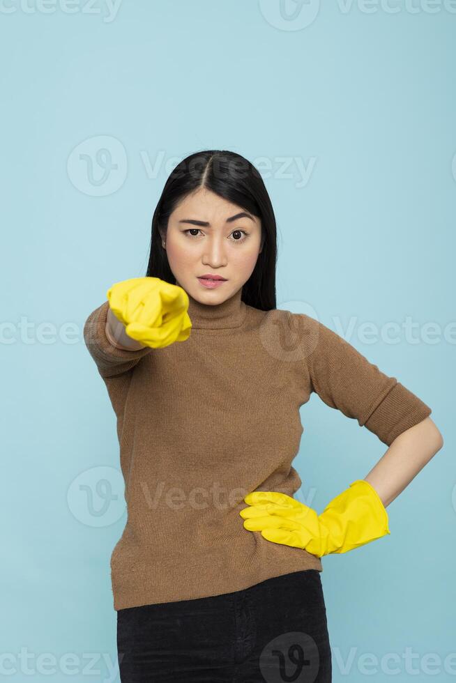 Tired female janitor poses in studio with blue background, pointing finger forward in frustration. Asian housekeeper wearing rubber gloves, gesturing at viewer with stern expression and raised eyebrow photo