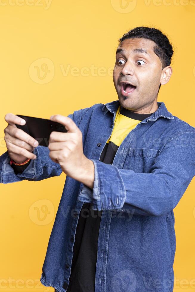 Happy joyous indian man playing online games against studio background, single player enjoying a competition tournament on mobile phone. Young adult with a cheerful expression. photo