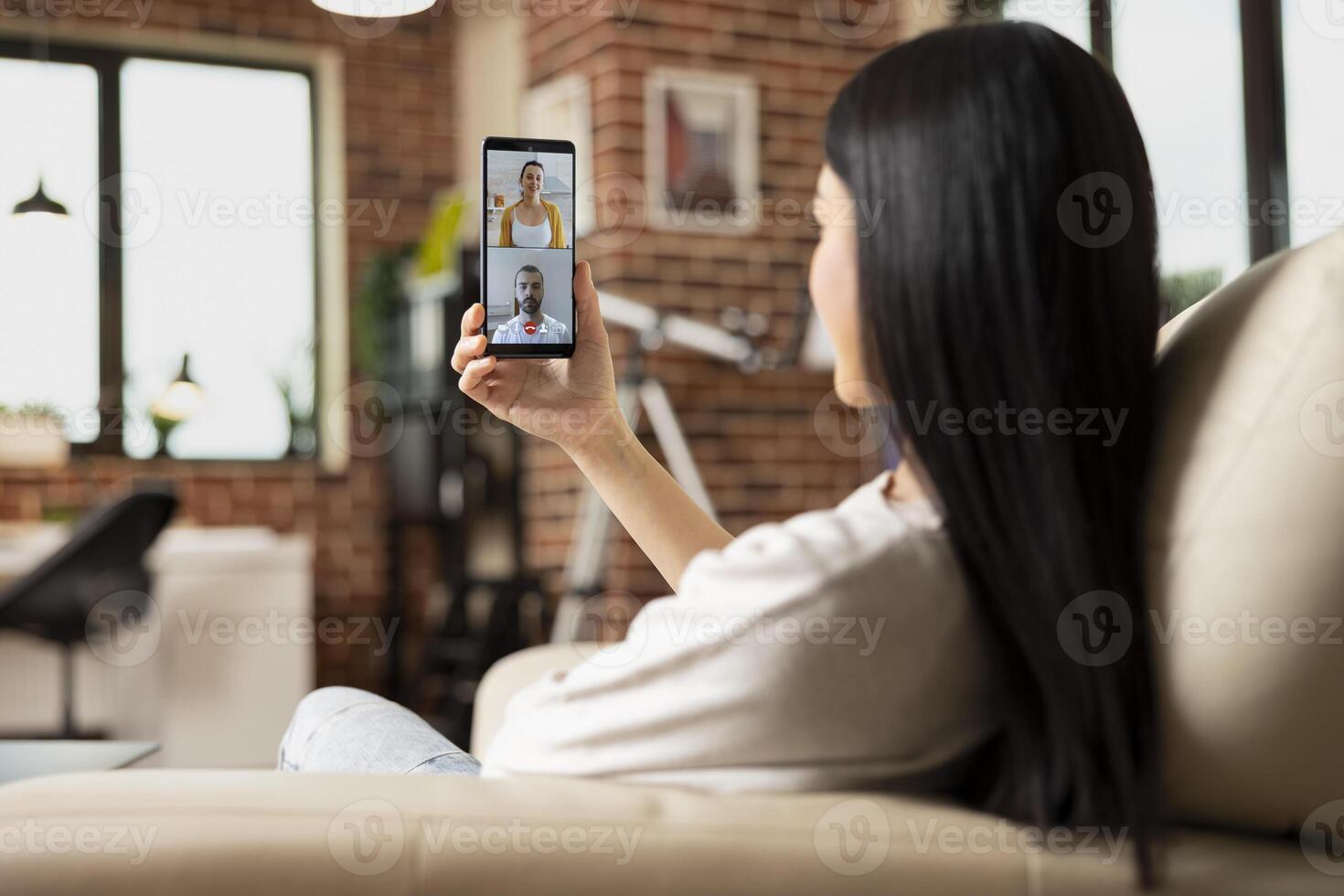 Asian businesswoman sitting on sofa with cellphone in hand, having conversation with long distance friends. Remote female manager talking with caucasian coworkers on call, sharing project ideas. photo