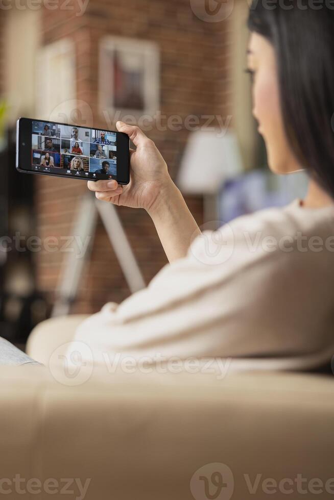 Back view of female employee on call with multiracial colleagues during online team briefing. Businesswoman seated on couch, attends virtual group meeting on mobile phone from home. photo