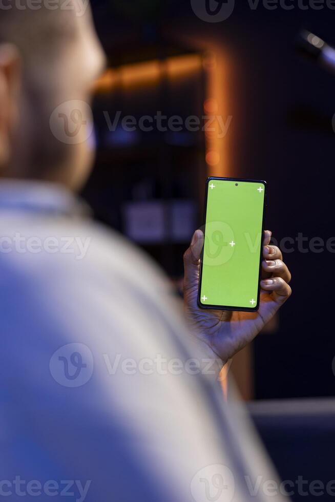 Black remote worker looking at green screen on his device at home, relaxing in the living room and solving his freelancing tasks. Young adult being casual in his trendy apartment, teleworking. photo