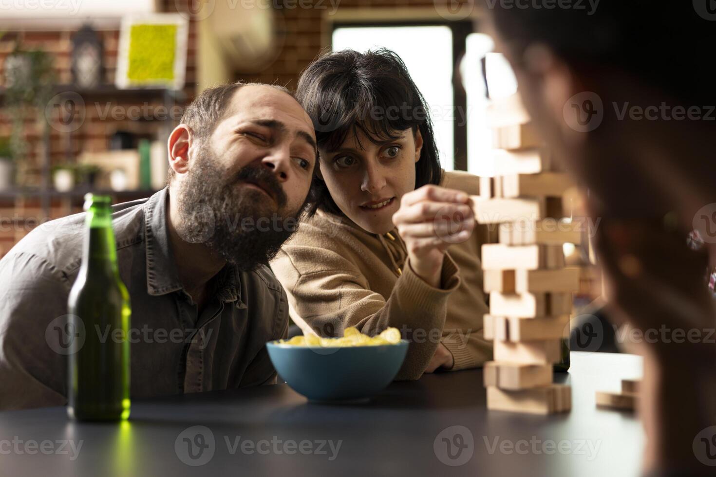 Young caucasian woman intensely focused on pulling wooden block from a jenga tower during tabletop game. Seated with snacks and drinks as her male friend watches the tense moment unfold. photo